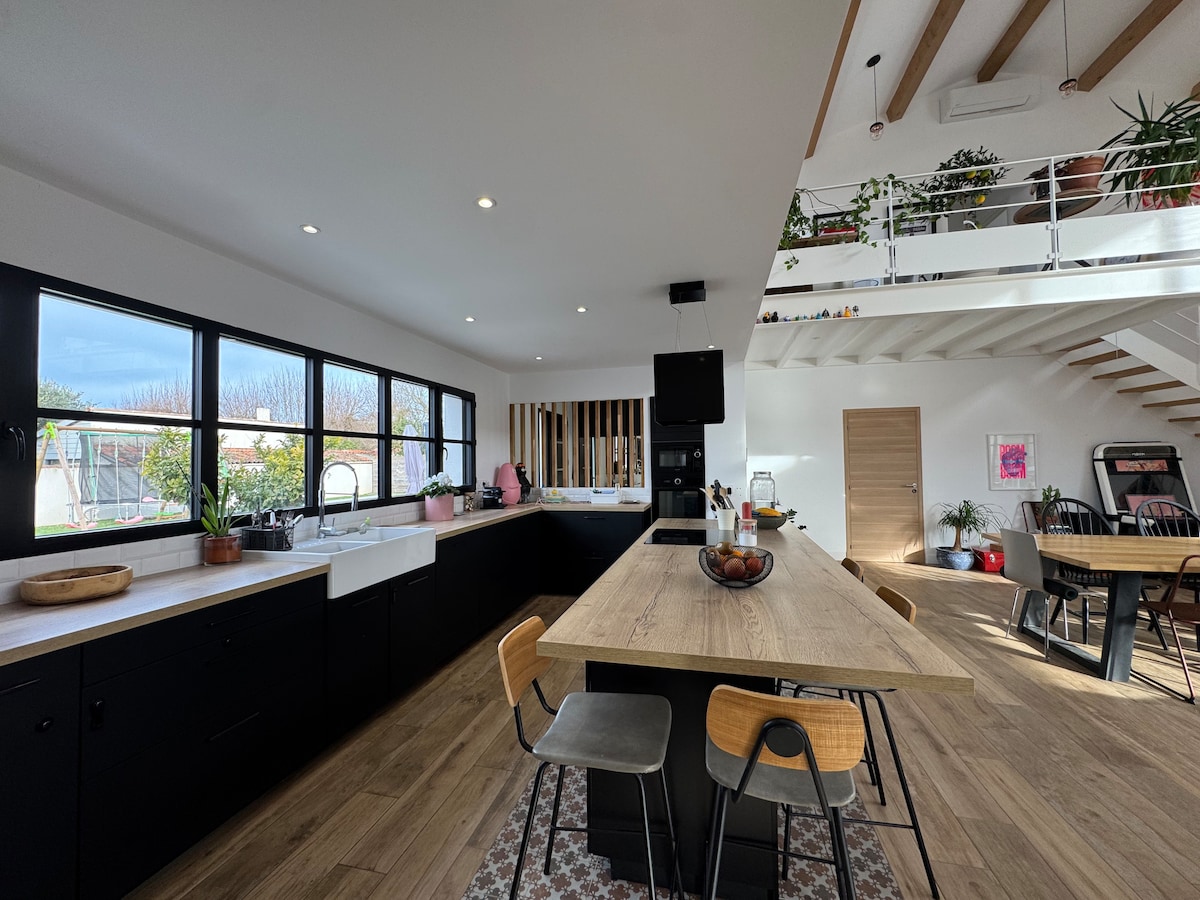 A modern kitchen features a large wooden island with seating for several people. Black cabinetry contrasts with white countertops. Expansive windows allow natural light to fill the space, showcasing a view of the outdoor area. Plants are placed thoughtfully around the room for a refreshing touch.