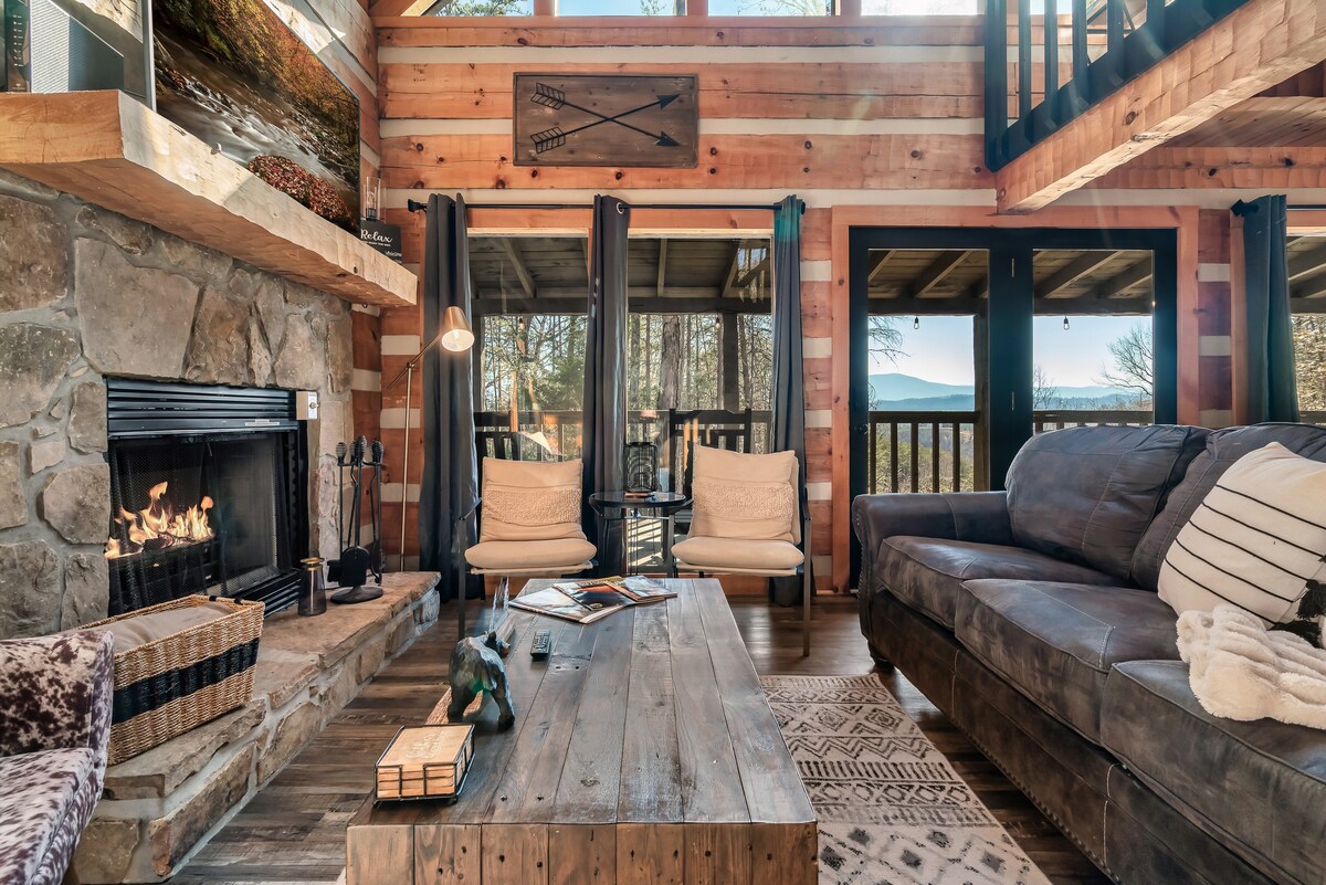 A spacious living area features a stone fireplace, with a warm fire flickering inside. Comfortable seating includes two light-colored armchairs and a large dark sofa. A wooden coffee table sits in the center, displaying magazines and decorative items. Large windows reveal natural light and mountain views.