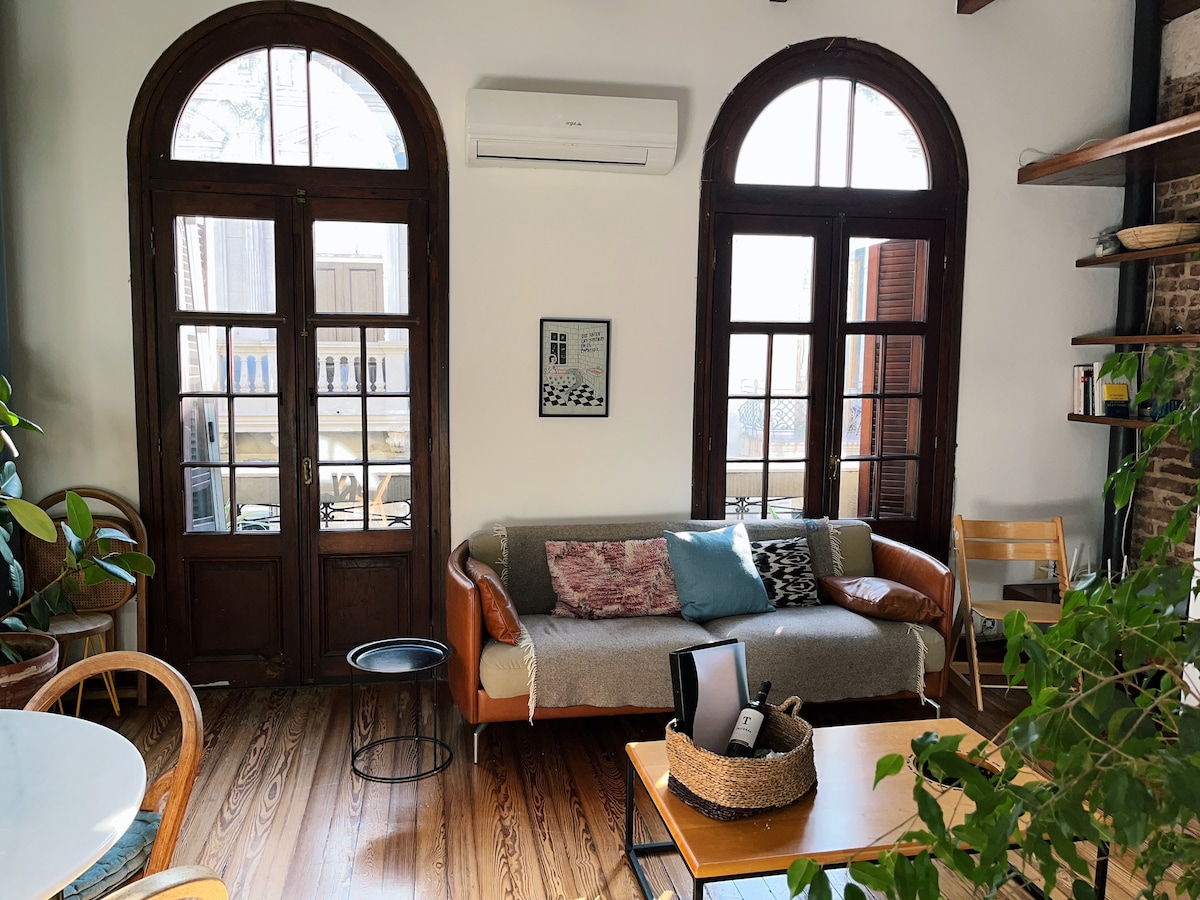 A bright and airy living area features large arched windows allowing natural light to fill the space. A cozy sofa is complemented by a mix of plants and decorative pillows. A wooden table and shelves add warmth, with a comfortable chair nearby.