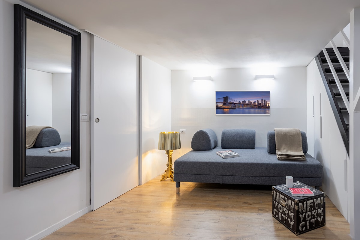 A cozy living area features a spacious gray sofa with light-colored throws positioned against a bright wall. A decorative coffee table with magazines sits on the light wood floor. A full-length mirror enhances the space, while soft lighting creates an inviting ambiance.