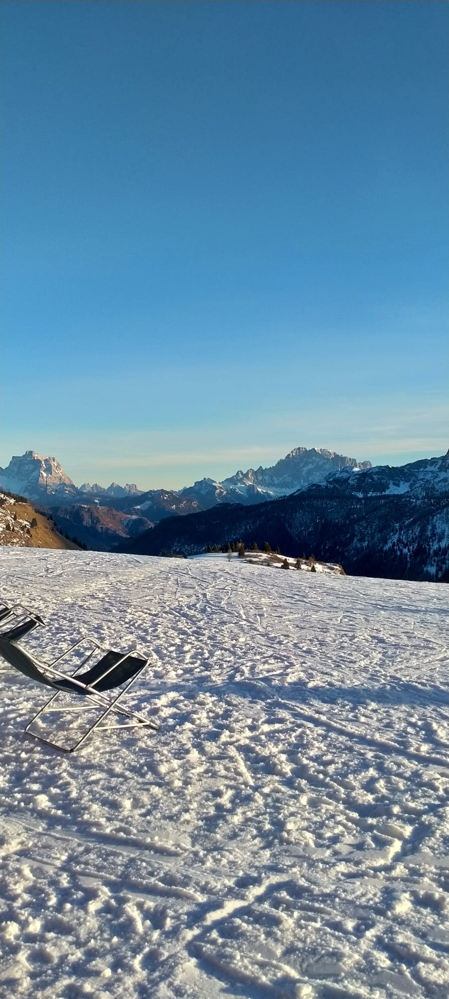 A snow-covered landscape stretches across the image, framed by majestic mountains in the distance. Two lounge chairs are positioned on the snow, inviting relaxation while taking in the panoramic views of the surrounding peaks and valley.