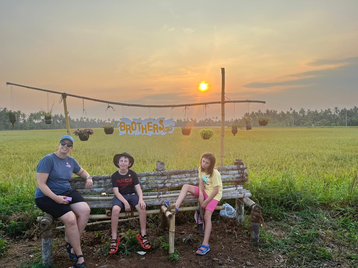 A wooden bench is positioned in a lush green field at sunset, with the sun setting on the horizon. Three figures, including children, are seated on the bench, enjoying the tranquil rural setting. Hanging planters add charm to the backdrop.