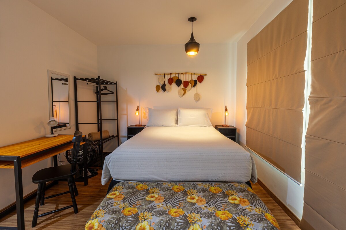 A spacious sleeping area is highlighted by a comfortable bed, accompanied by a decorative throw. A small desk and chair are situated nearby, providing a workspace. Soft lighting is provided by lamps on each side of the bed, with a large window partially covered by modern blinds.