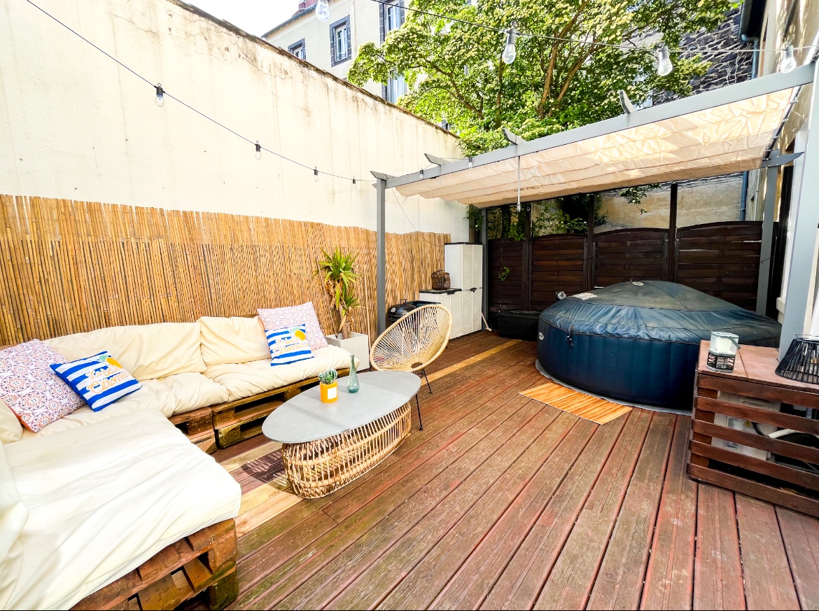 An outdoor terrace features a cozy seating area furnished with a light-colored sofa and a round coffee table. A shaded section is provided by a canopy over a jacuzzi, which is surrounded by wooden decking and greenery, creating a tranquil setting.
