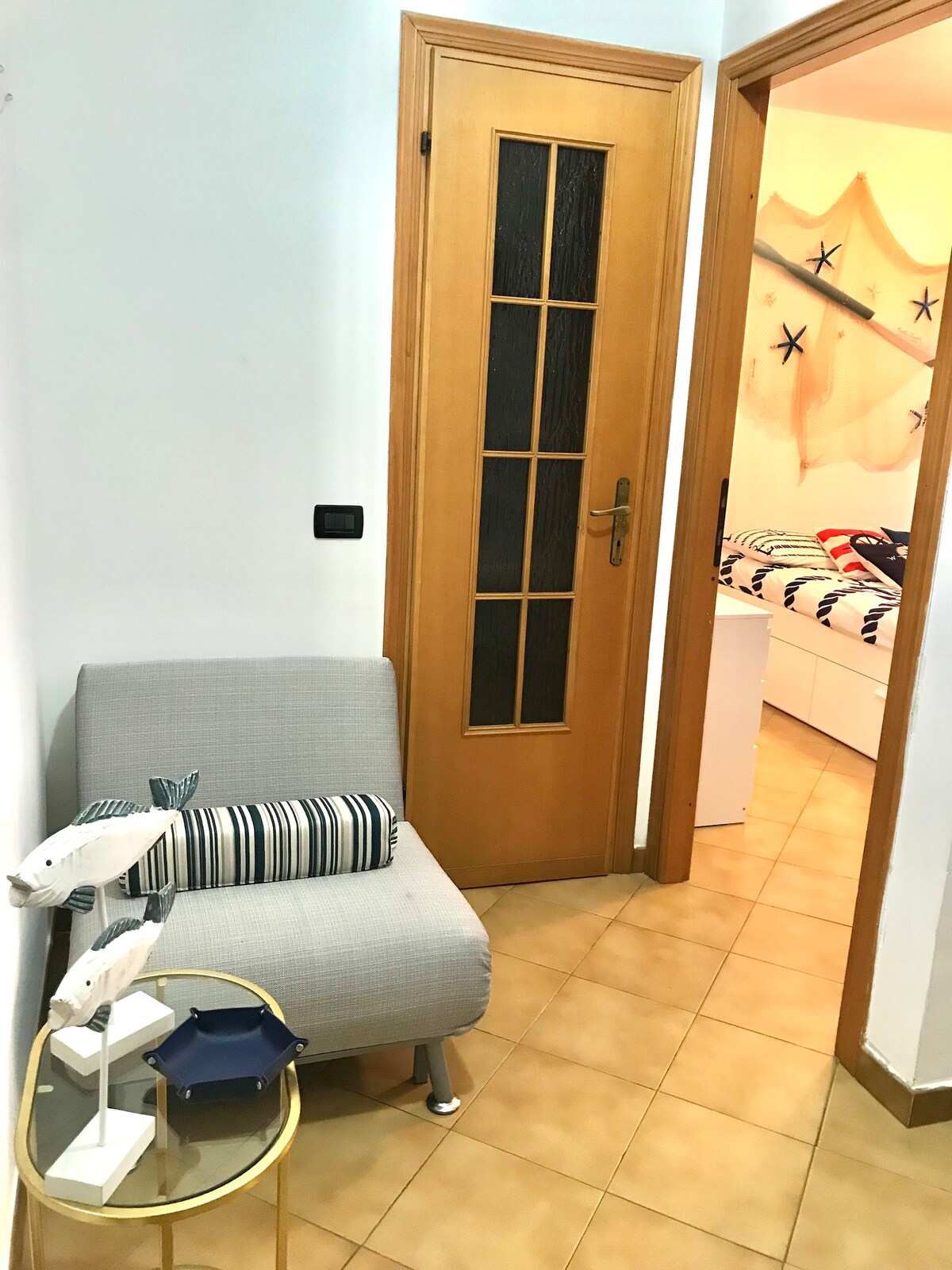 A small entryway features a light-colored chair with striped cushions and a round side table. A wooden door with glass panels leads to another room. The floor is tiled in warm tones, and nautical-themed decor is visible in the adjacent room.