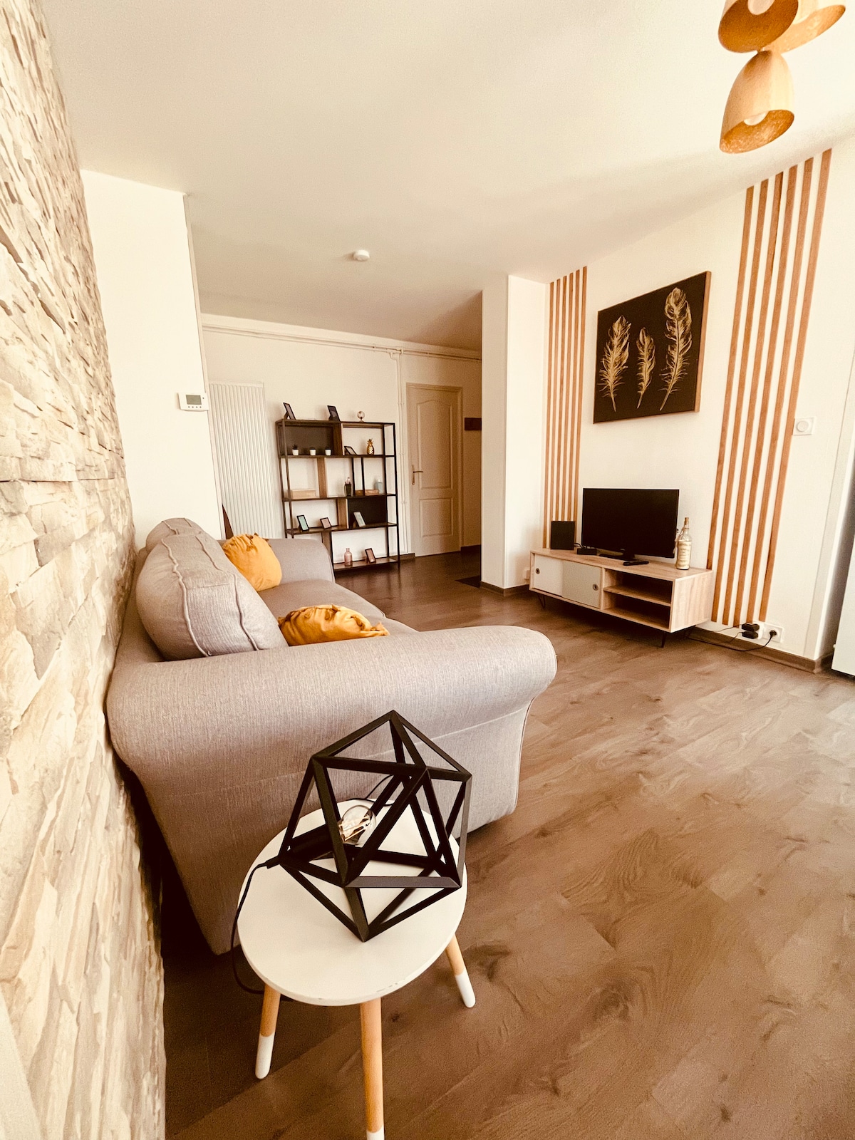 A well-lit living area features a comfortable gray sofa adorned with decorative cushions. A small white side table with a geometric lamp sits nearby. Opposite, a media unit displays a television, with a sleek shelf in the background showcasing books and decorative items.