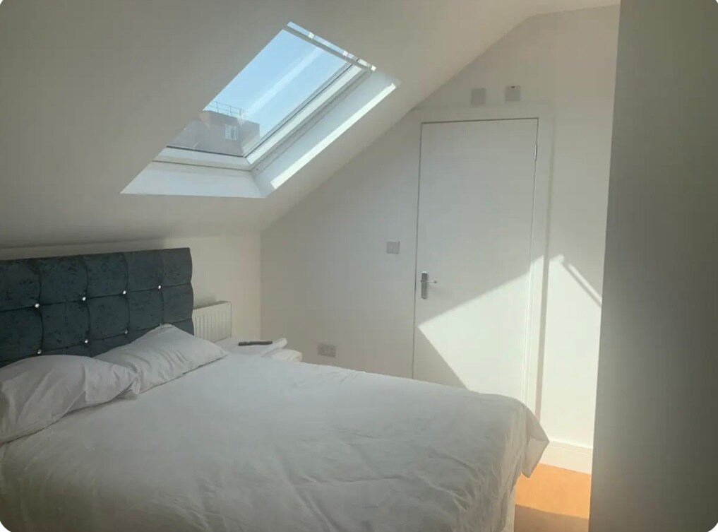 A well-lit bedroom showcases a spacious double bed dressed in white linens. A skylight above allows natural light to illuminate the room, creating a bright atmosphere. A door can be seen on one side, leading to a private area.