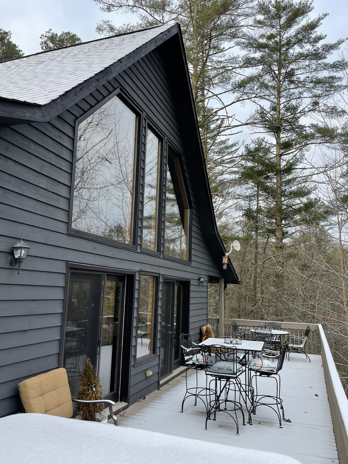 The cabin’s exterior features large windows that provide plenty of natural light. A spacious deck is furnished with metal tables and chairs, offering ample seating for outdoor enjoyment. The surrounding trees and elevated design create a serene atmosphere.