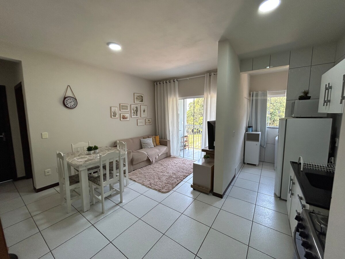 A light-filled living space features a cozy seating area with a sofa, a dining table set for four, and a well-equipped kitchen. Large windows provide a view of the outdoors, while soft curtains diffuse natural light, creating an inviting atmosphere for relaxation and dining.