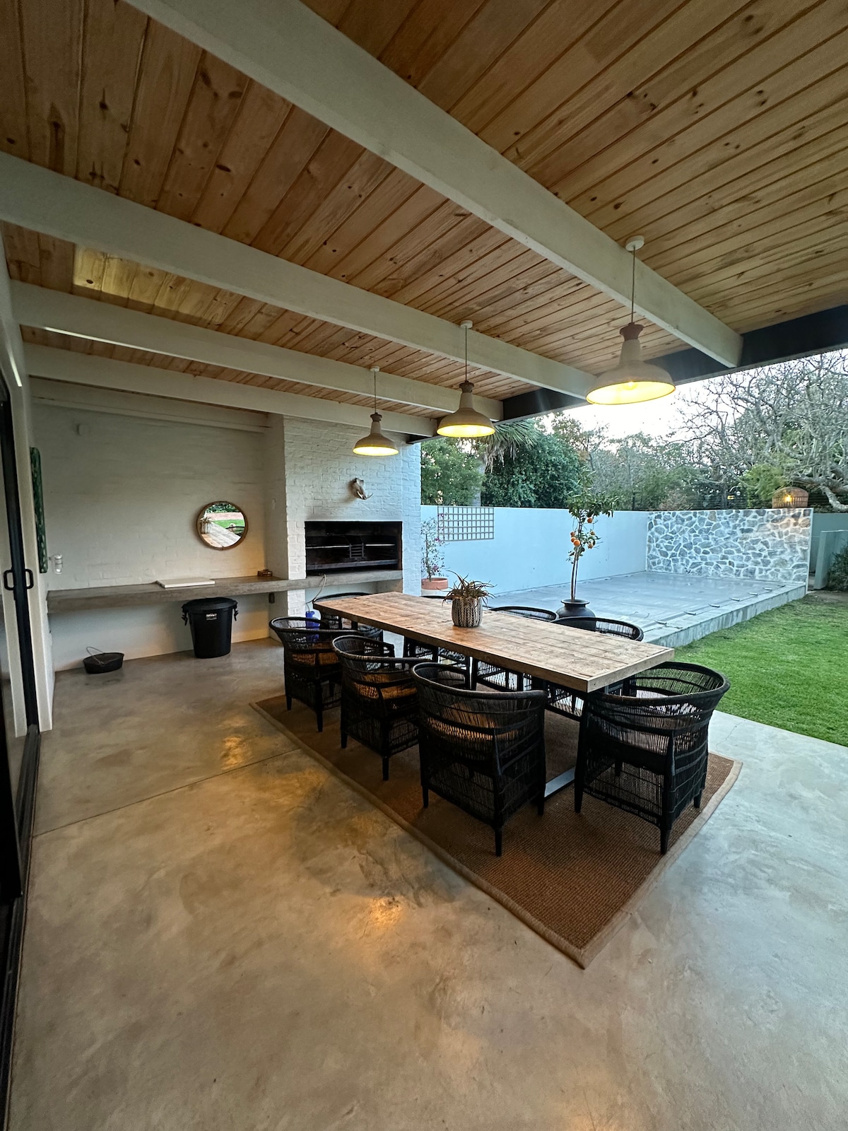 An outdoor dining area is presented under a wooden ceiling with exposed beams. A long wooden table, surrounded by black woven chairs, offers seating for several guests. A built-in braai and decorative mirror are visible, with a spacious garden area beyond.