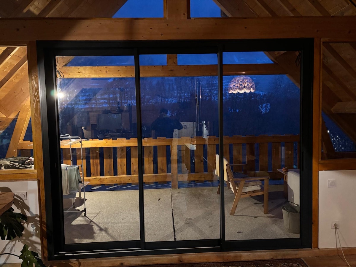 A large sliding glass door leads to a terrace, showcasing wooden railings and an open view of the surrounding nature. Soft lighting from an overhead fixture enhances the space, which features a sloped wooden ceiling and a comfortable chair positioned on a gray floor.