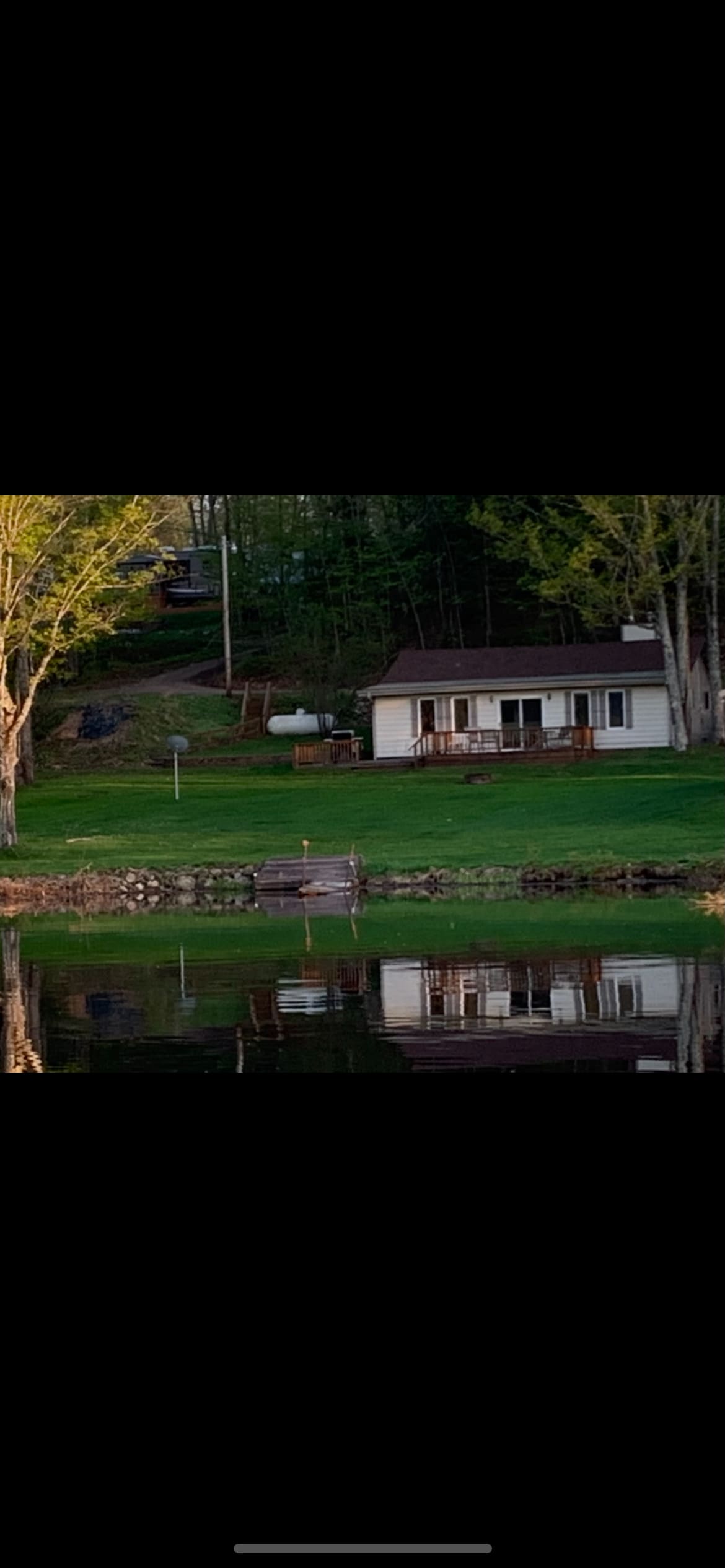 A cabin is situated beside a tranquil lake, with green grass surrounding it. The structure features a porch with railing and large windows, reflecting softly on the water's surface. Lush trees can be seen in the background, contributing to the serene environment.