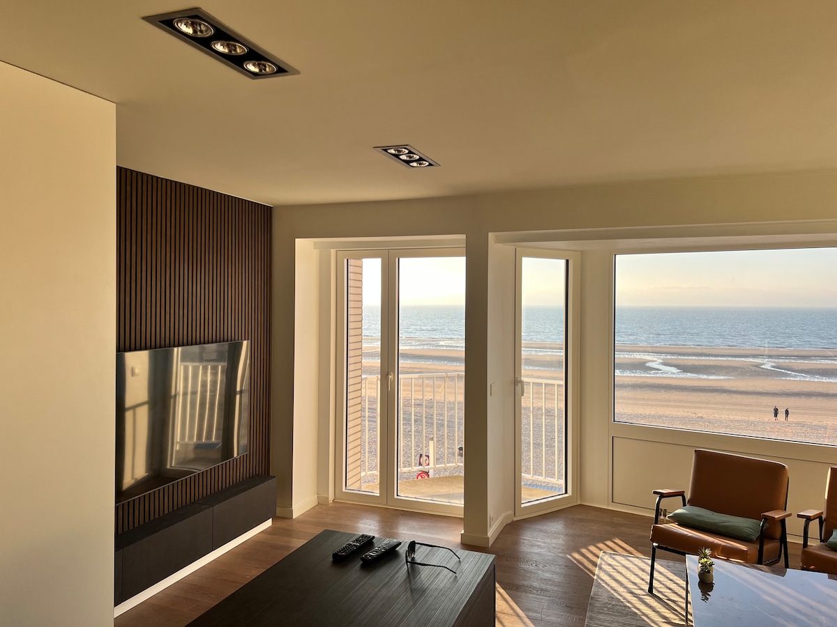 A spacious living area is complemented by large windows framing a broad seaview. Natural light illuminates the space, highlighting a modern flat-screen television mounted on a wood-paneled wall. A comfortable green armchair and minimalist coffee table enhance the inviting ambiance.