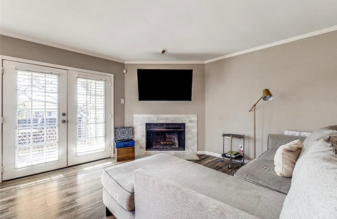 A comfortable living area is displayed, featuring a sectional sofa and a modern coffee table. A gas fireplace is positioned beneath a wall-mounted television. Natural light enters through double doors that lead to a balcony, complemented by neutral-toned walls and hardwood flooring.