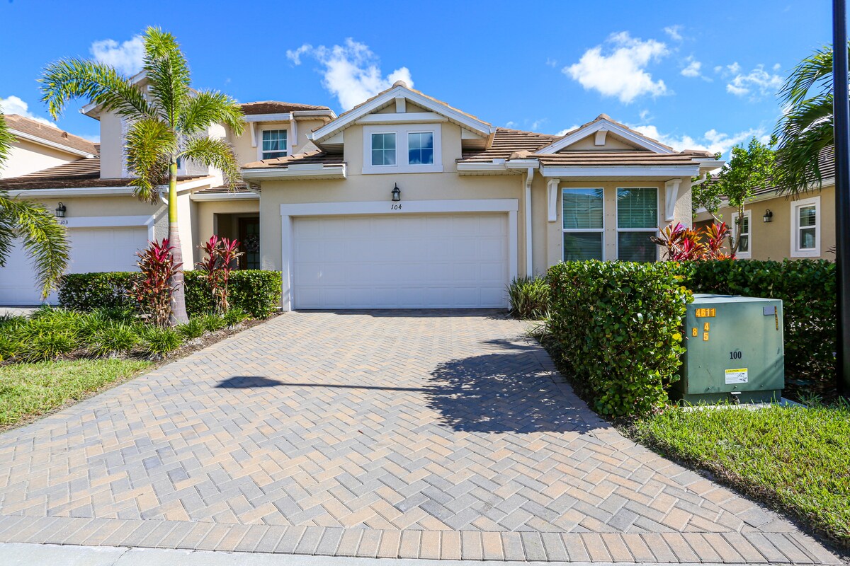 A single-level townhouse is showcased with a spacious driveway made of patterned pavers. Lush greenery frames the entrance, and palm trees are visible in the landscaping. The front features a large garage door and several windows allowing natural light to enter.