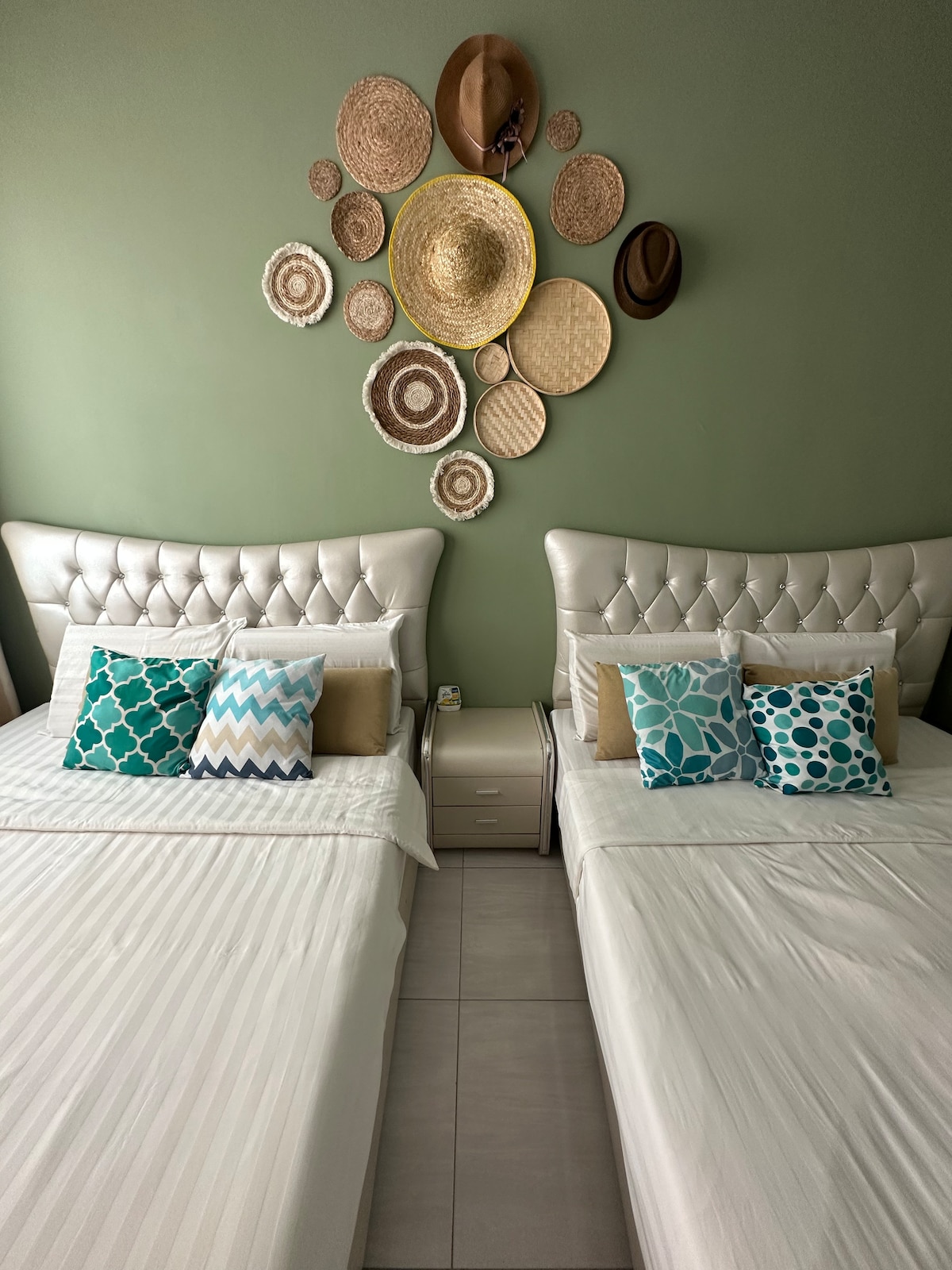 Two spacious beds are positioned in the room, each adorned with a colorful array of pillows. A decorative arrangement of woven wall art and hats enhances the ambiance. The calming green walls provide a soothing backdrop, complemented by modern bedside tables.