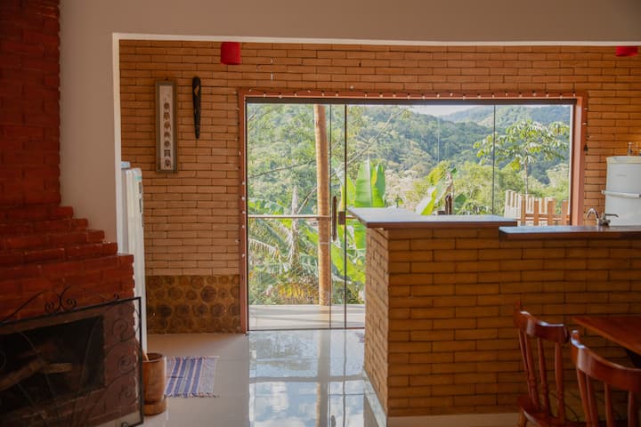 Integrirani dnevni boravak i kuhinja s izloženom ciglom i obiljem prirodnog svjetla. Staklena vrata vode na terasu s pogledom na šumu, stvarajući ugodan, jednostavan prostor povezan s prirodom.