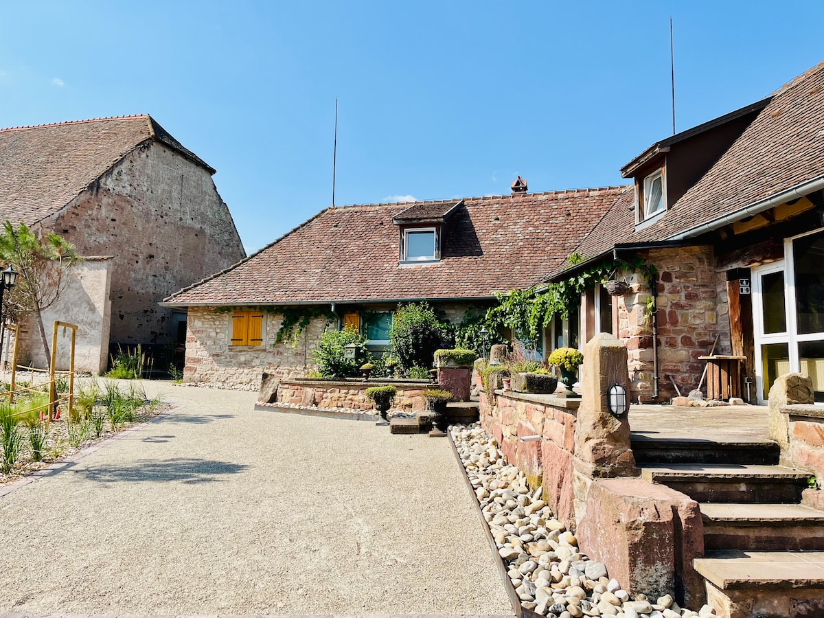 The exterior of the property showcases a historic building with stone walls and a sloped roof, surrounded by a gravel courtyard. Lush greenery adds a natural touch, and an inviting pathway leads to the entrance, creating a charming and serene atmosphere.