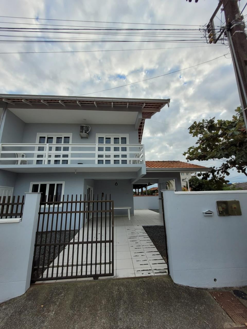 The exterior of the house is captured, featuring a light blue facade with a balcony on the second floor. A gated entrance leads to a spacious outdoor area with a stone pathway and a few potted plants.