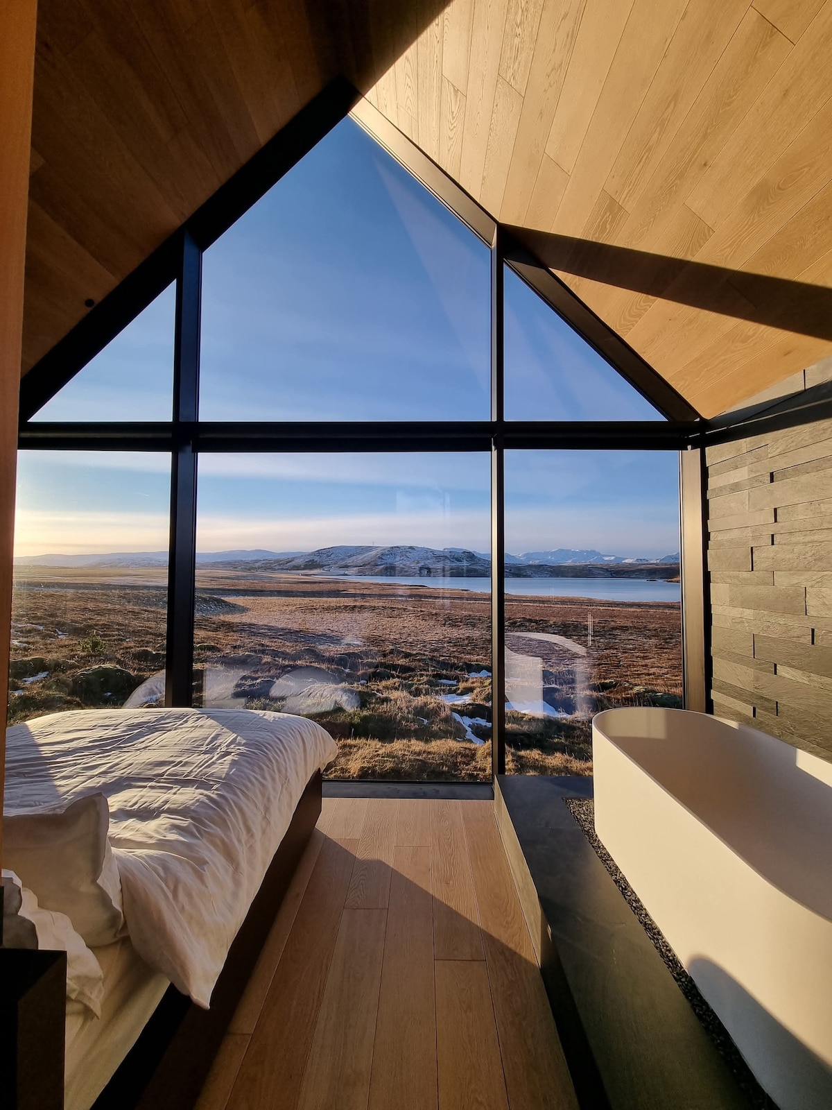 A cozy bedroom features large floor-to-ceiling windows providing a panoramic view of the surrounding landscape. A spacious bed is positioned near the windows, while a modern freestanding tub complements the minimalist design, creating a serene atmosphere.