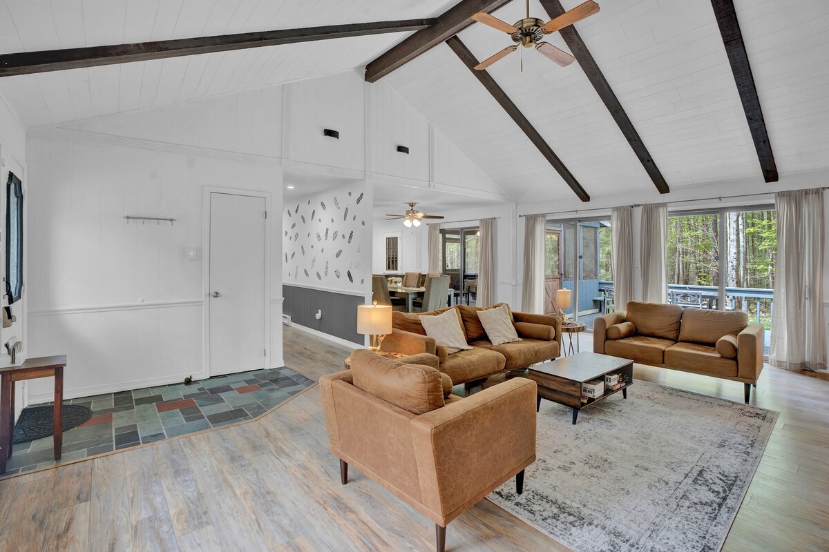 A spacious living area features high, exposed beam ceilings and large windows, allowing for natural light. Comfortable seating is arranged around a central coffee table, and soft lighting from nearby lamps enhances the cozy ambiance. A dining area can be seen in the background, with a view of outdoor greenery.