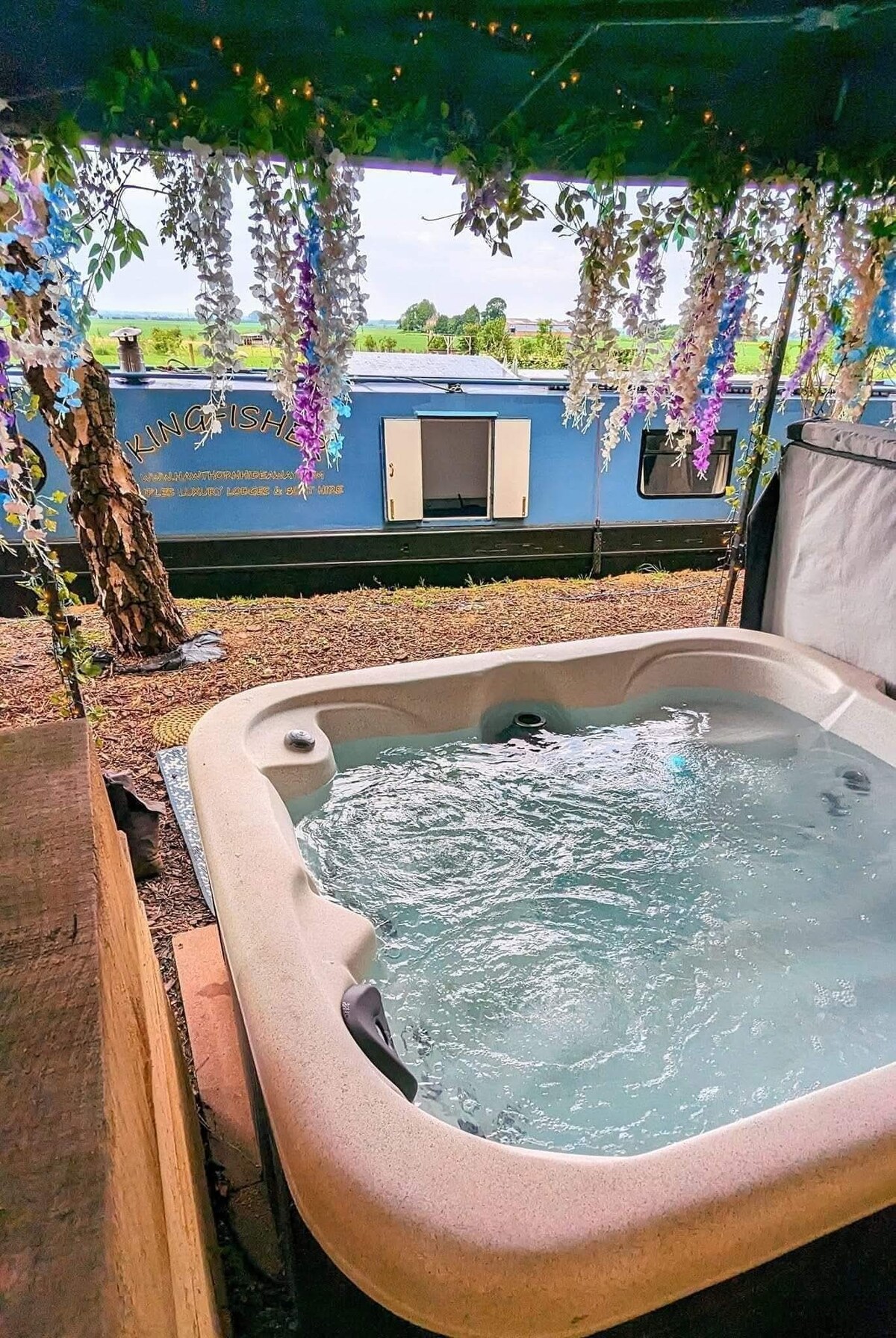 A covered hot tub is positioned under a canopy adorned with hanging flowers, offering a relaxing view. The clear water ripples gently, while a blue canal boat can be seen in the background against a serene landscape.