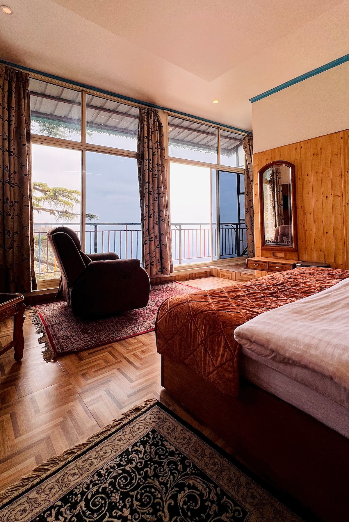 A serene bedroom features a spacious bed with warm bedding, accompanied by a comfortable armchair. Large windows offer an expansive view of the valley, framed by tall curtains. Elegant wooden accents and a decorative rug enhance the cozy atmosphere.