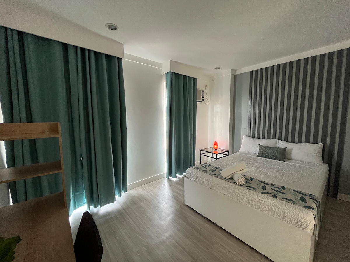 A well-lit room features large windows draped with teal curtains, allowing natural light to enter. An inviting bed positioned against a striped accent wall is accompanied by a small bedside table with a lamp. A wooden desk and chair are also included in the space.
