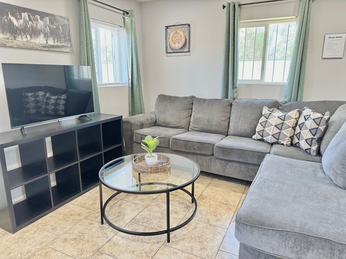 The living room is furnished with a large sectional sofa and a circular glass coffee table. A Smart TV is positioned on a media unit along one wall, with a piece of wall art featuring horses above it. Natural light enters through two windows with light curtains.