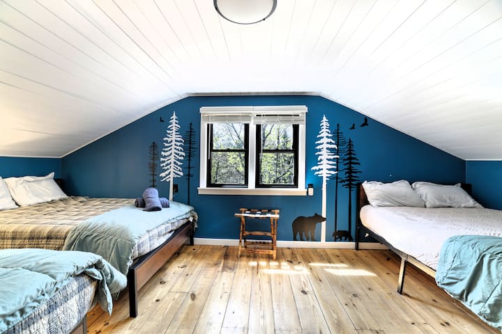 Arriba, dormitorio loft soleado con 3 camas tamaño queen, mural de bosque y cálidos pisos de madera