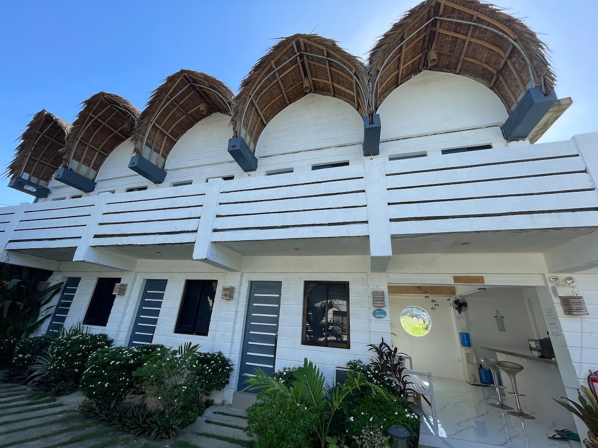 A modern building features a series of arched roofs with thatched accents, providing character to the design. The exterior is painted white, with multiple windows framed in dark colors. Lush greenery and flowers surround the area, adding to the inviting ambiance.