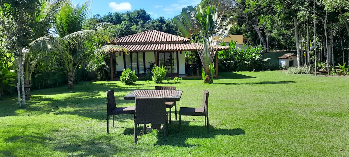 A spacious outdoor area features a grassy lawn surrounded by tropical plants. In the foreground, a four-seat dining table is positioned on the grass, with a view of the house, which has a tiled roof and large glass doors opening into the garden.