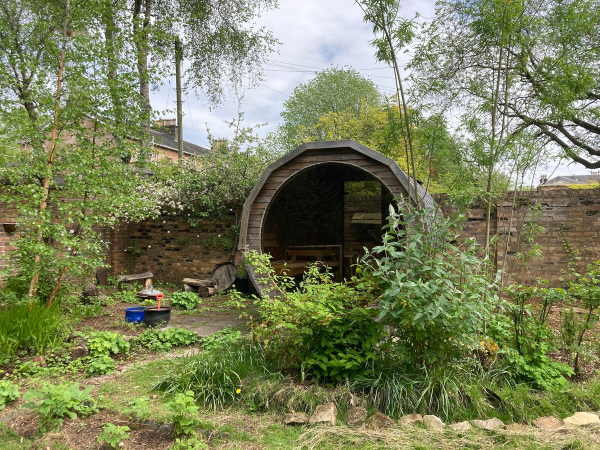 A wooden structure resembling a cozy, circular cabin is nestled within a lush garden. Surrounded by greenery, it features a table and benches inside, inviting relaxation. The scene includes various plants, trees, and a stone pathway leading to this charming outdoor space.