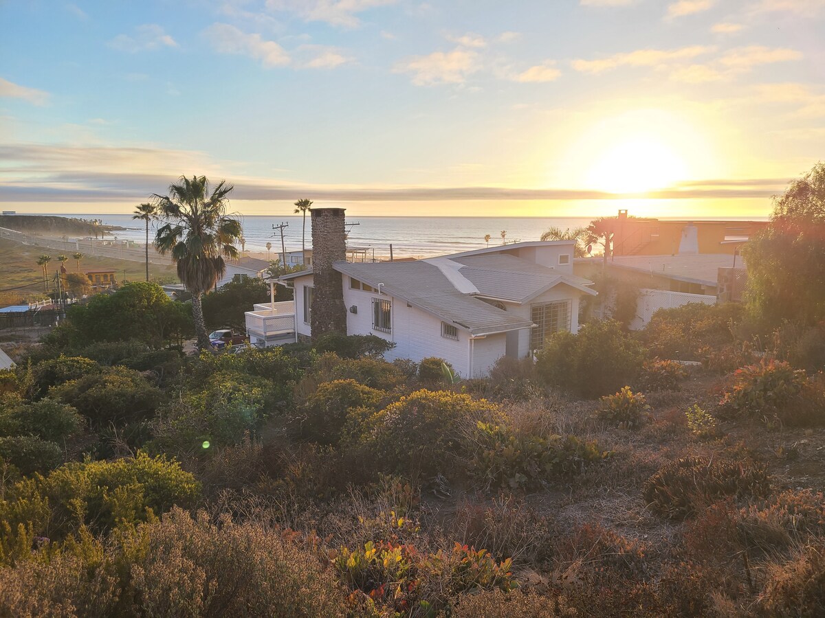 A beach house is set among lush landscaping, with a stone chimney visible. The sun rises in the background, casting a warm glow over the scene. The ocean is seen in the distance, framed by palm trees and coastal vegetation.