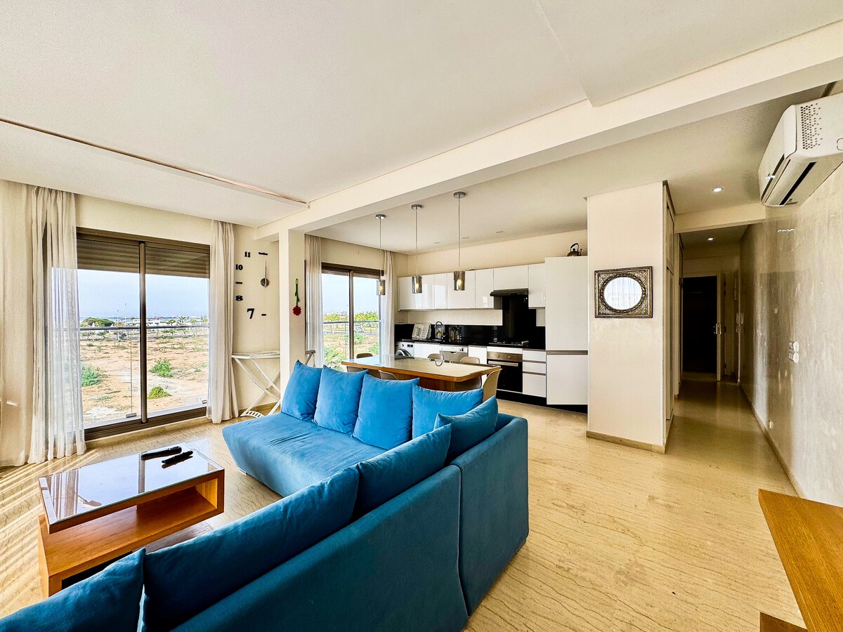 A spacious living area features light-colored walls and a comfortable blue sofa facing large windows that provide a view of the outdoor landscape. The open kitchen, equipped with modern appliances, is visible in the background, enhancing the room's airy feel.