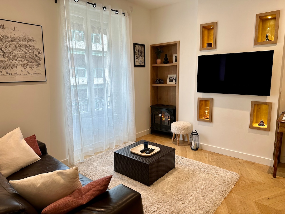 A cozy living area features a dark leather sofa with decorative pillows and a soft area rug. A modern coffee table is placed centrally, while a television is mounted on the wall above a wooden unit with illuminated shelves. Natural light filters through sheer curtains covering a large window.