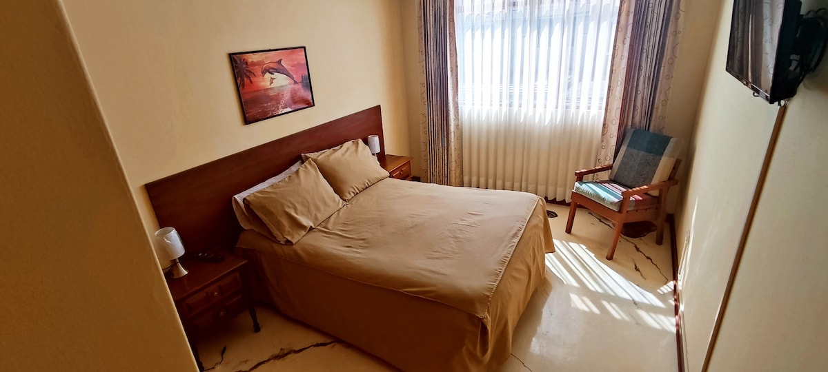 A serene sleeping area features a spacious bed with neutral-toned bedding. Natural light filters through sheer curtains, illuminating the room. A comfortable chair sits in one corner, accompanied by a small side table. Wall art adds a subtle decorative element.