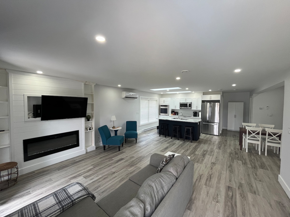 A spacious living area features comfortable seating with two blue chairs and a gray sofa. A modern fireplace is integrated into a white wall. The open-plan design connects to a well-equipped kitchen with stainless steel appliances and dining space with white chairs and a table.