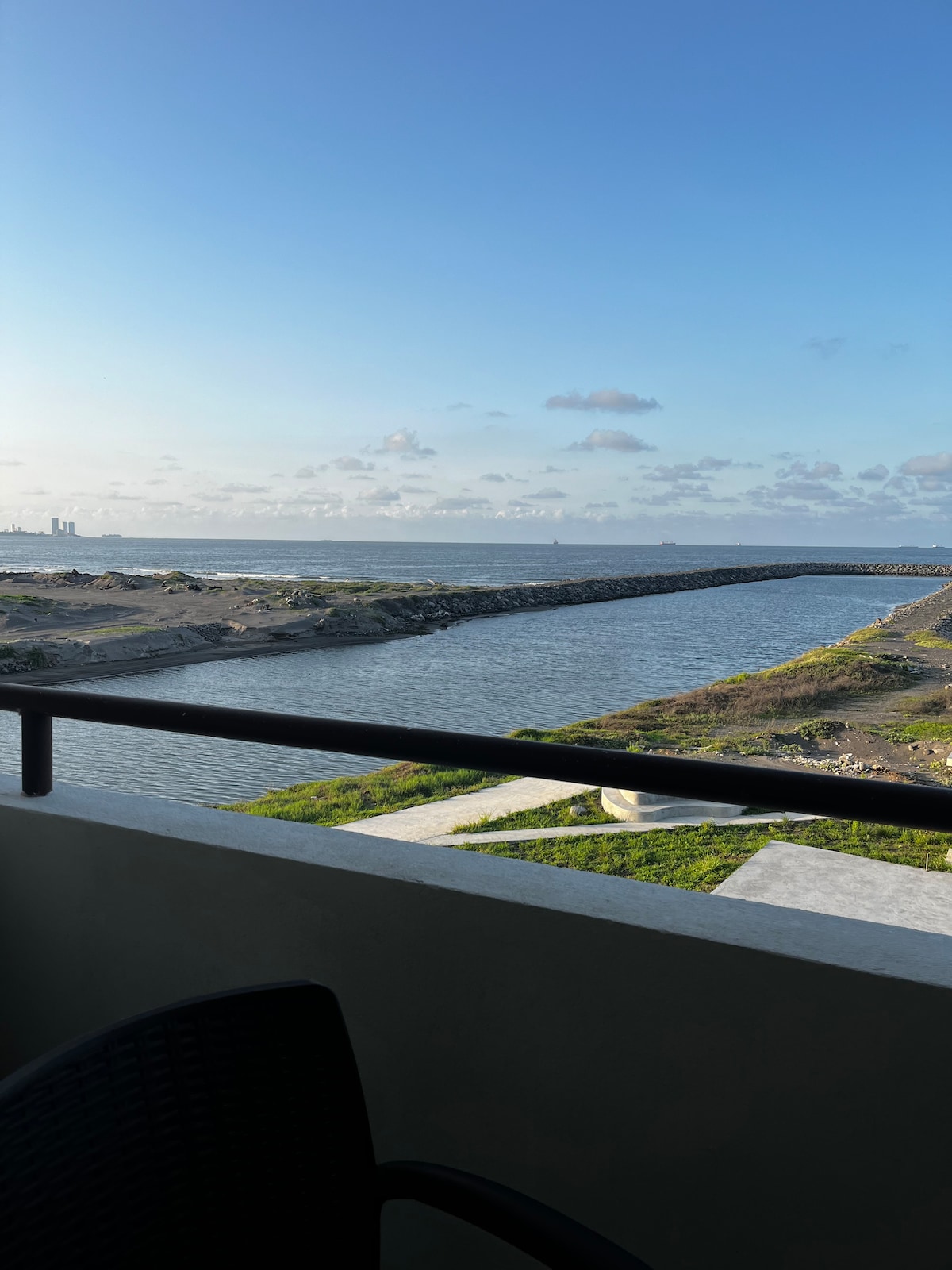 A scenic view from a balcony reveals a tranquil waterway, bordered by grassy banks. The expansive ocean is visible in the distance, complemented by a clear sky with soft clouds. A stone structure extends into the water, creating a natural frame for the picturesque landscape.