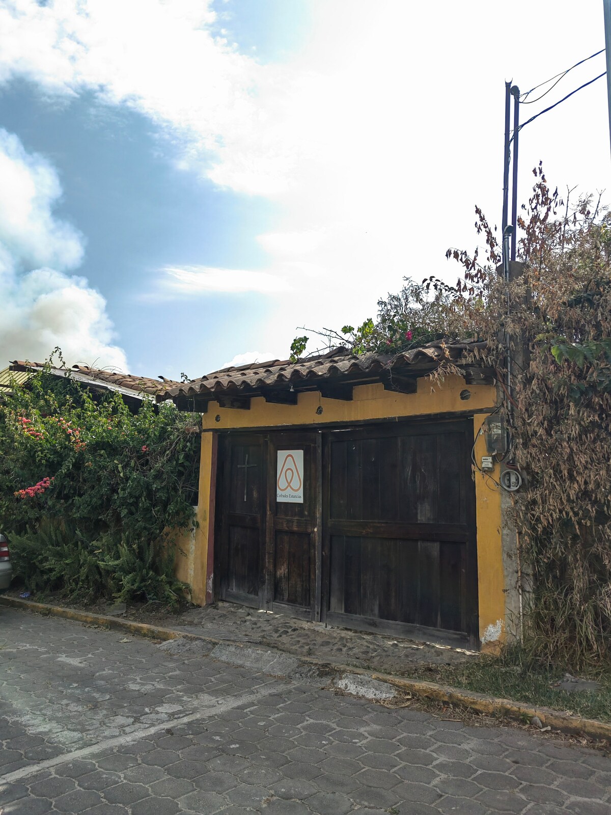 A warm, inviting entrance is framed by lush greenery and blooming plants. The rustic wooden gates, adorned with an Airbnb logo, lead to a charming property. Above, a clear blue sky and soft clouds create a serene backdrop, enhancing the welcoming ambiance.