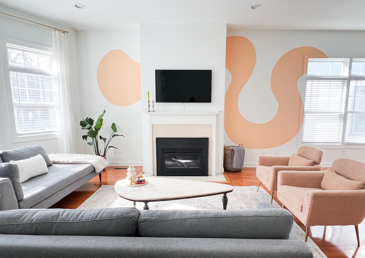 A spacious living area features soft gray sofas and two light orange accent chairs, arranged around a modern coffee table. A decorative fireplace serves as a focal point beneath a wall-mounted television. Natural light filters through large windows, illuminating the minimalist decor.