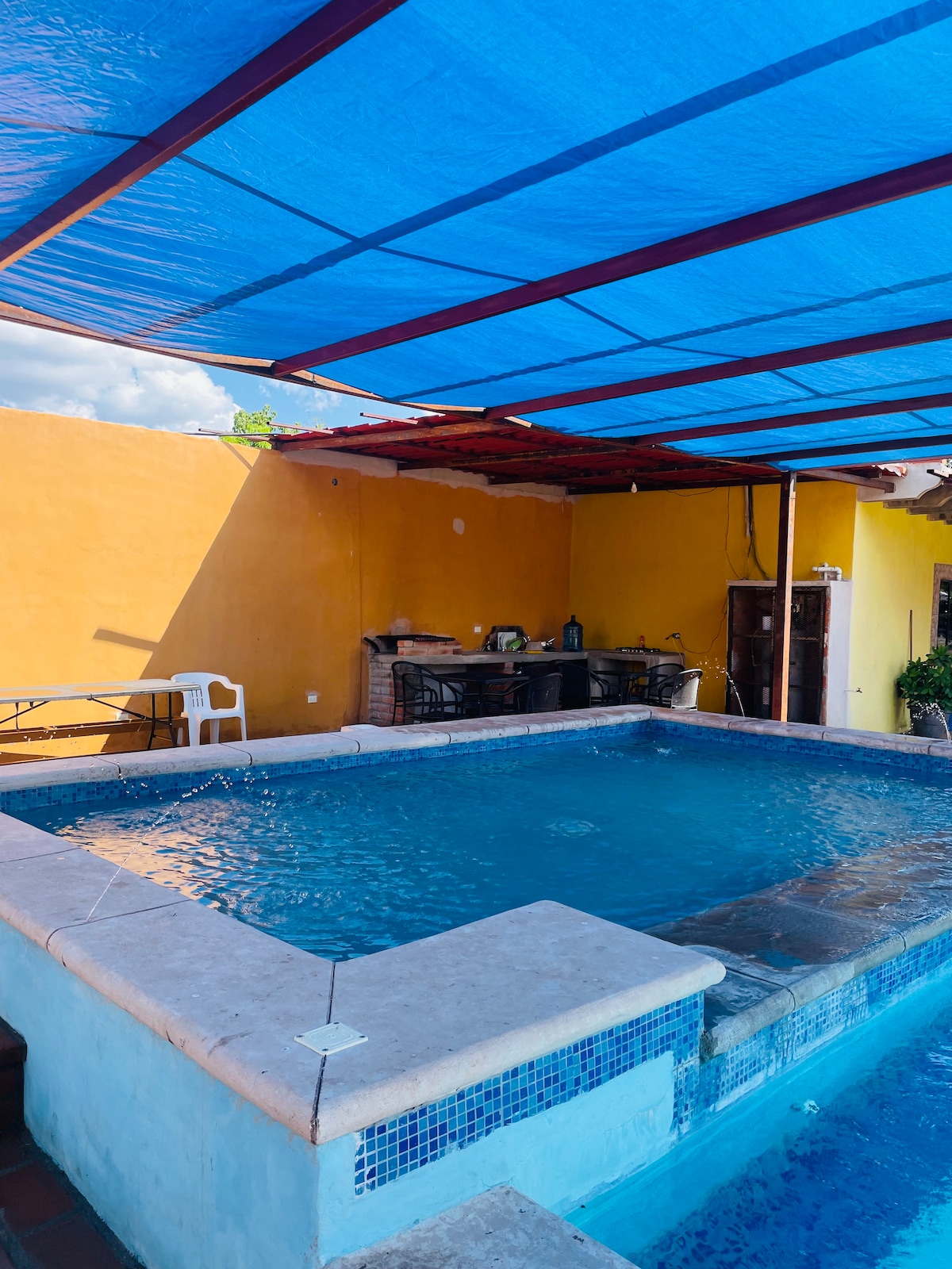A swimming pool is surrounded by a stone wall, featuring a light blue covering overhead for shade. The yellow wall in the background adds color, while an outdoor kitchen area with a barbecue grill and dining seating are visible nearby.