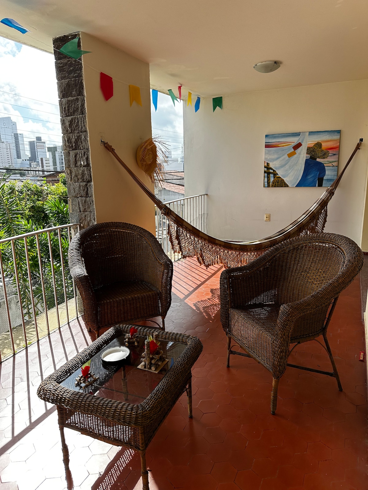A cozy balcony area is presented with two wicker chairs facing a small table. A colorful hammock stretches across the space, and a vibrant painting adorns the wall. Decorative bunting adds a festive touch to the outdoor setting, with greenery visible in the background.