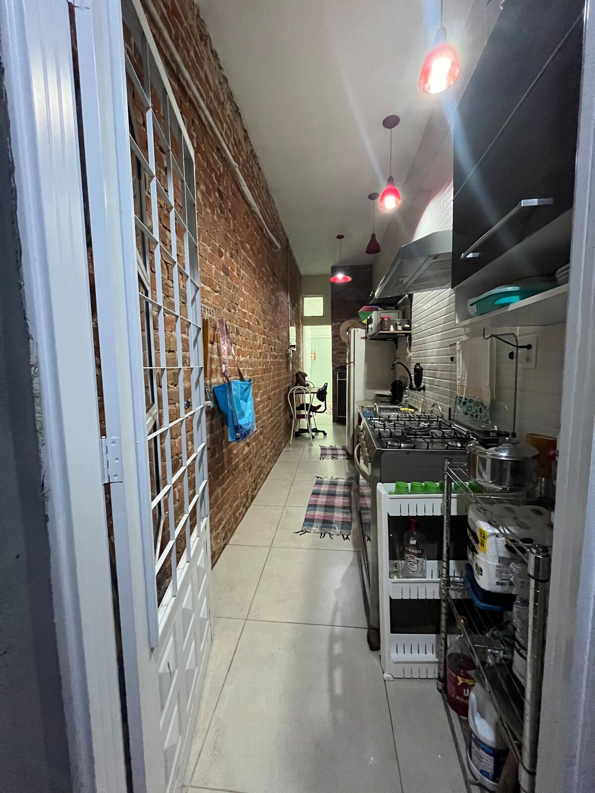 A narrow kitchen corridor features exposed brick walls and modern lighting. The space includes a cooking area with a stovetop and various kitchen utensils arranged on nearby shelves. A small mat lies on the tiled floor, adding a touch of comfort.