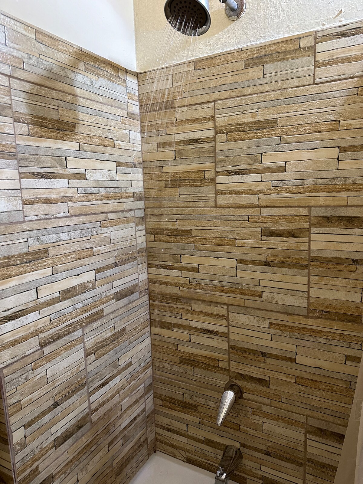 A modern shower area features textured stone walls in warm tones. Water flows from a sleek showerhead above, and a silver faucet is positioned on the wall below. The space is well-lit, highlighting the contemporary design.