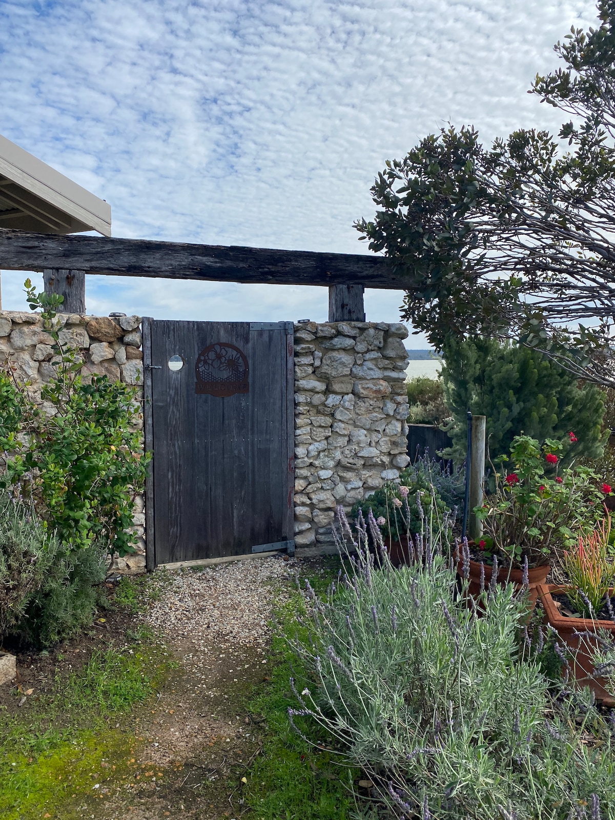 An inviting pathway leads to a rustic wooden gate framed by a stone wall. Lush greenery and colorful flowers, including lavender, are scattered throughout the garden, creating a serene outdoor entrance.