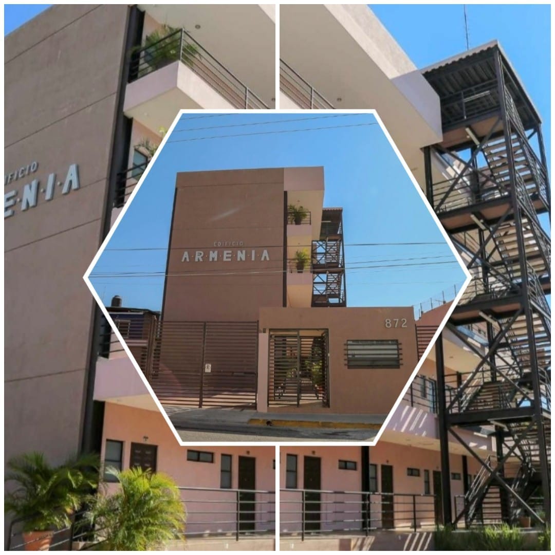 The exterior of a modern building named 'Edificio Armenia' is depicted, featuring a combination of warm-colored walls and metal accents. A secure entrance with gates is visible, complemented by a staircase that leads to upper levels. Potted plants add a touch of greenery to the environment.