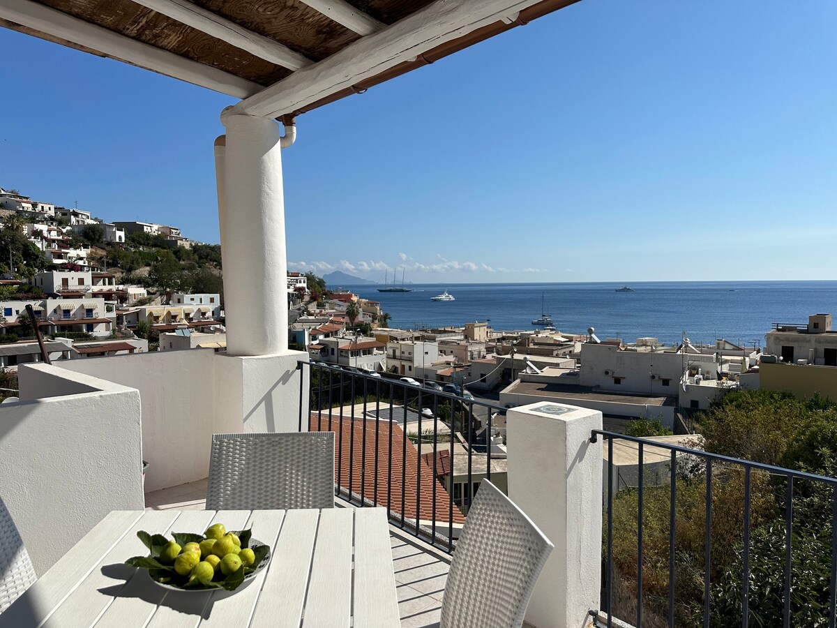 A terrace is featured with a white table surrounded by four chairs. A bowl of lemons sits at the center. In the background, a stunning view is visible, showcasing the sea and nearby coastal town with boats anchored in the water under a clear blue sky.