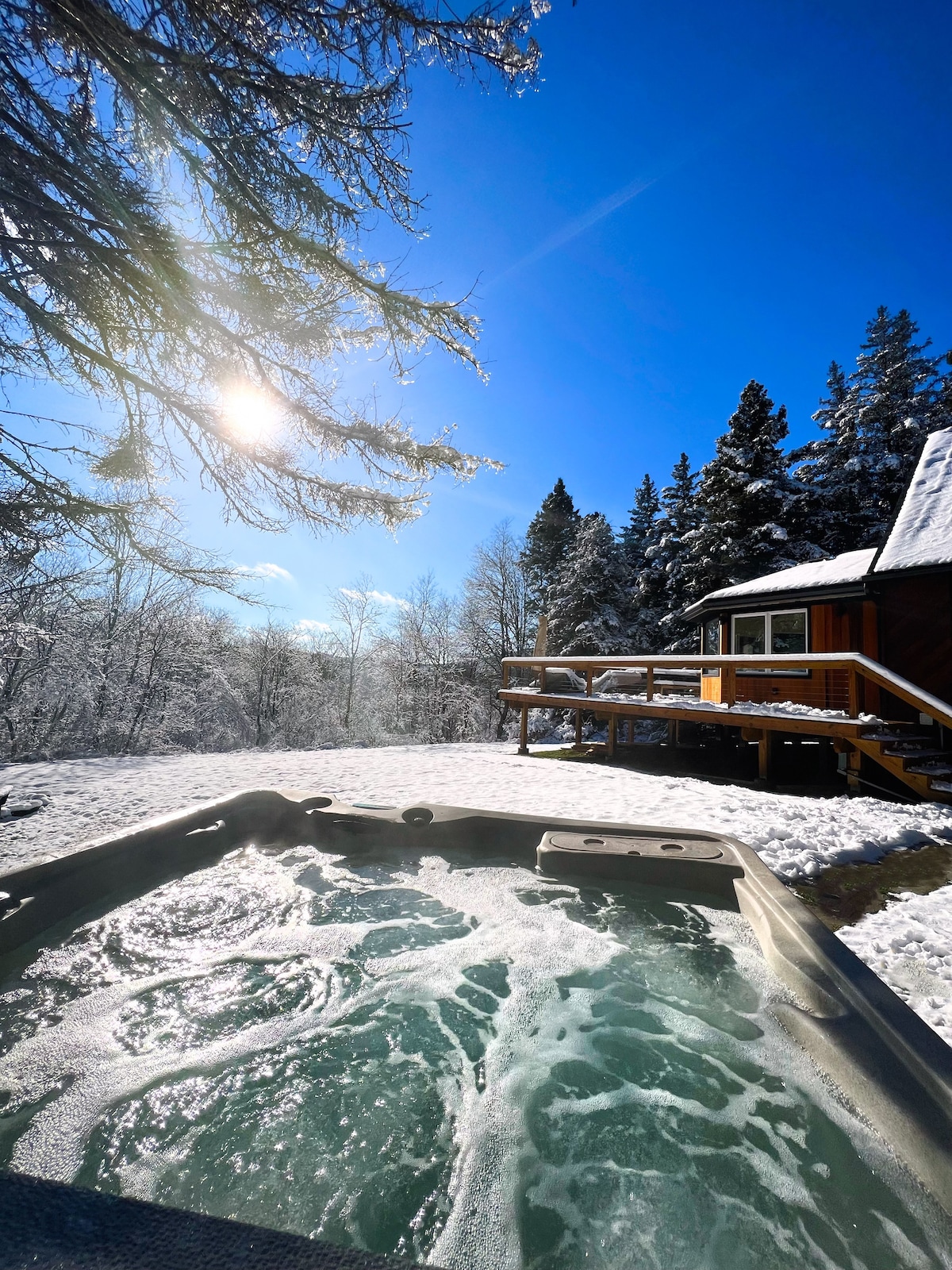 A hot tub is surrounded by snow-covered ground, with sunlight reflecting off the bubbling water. A wooden deck is visible in the background, framed by tall trees and a clear blue sky, providing a serene outdoor retreat.