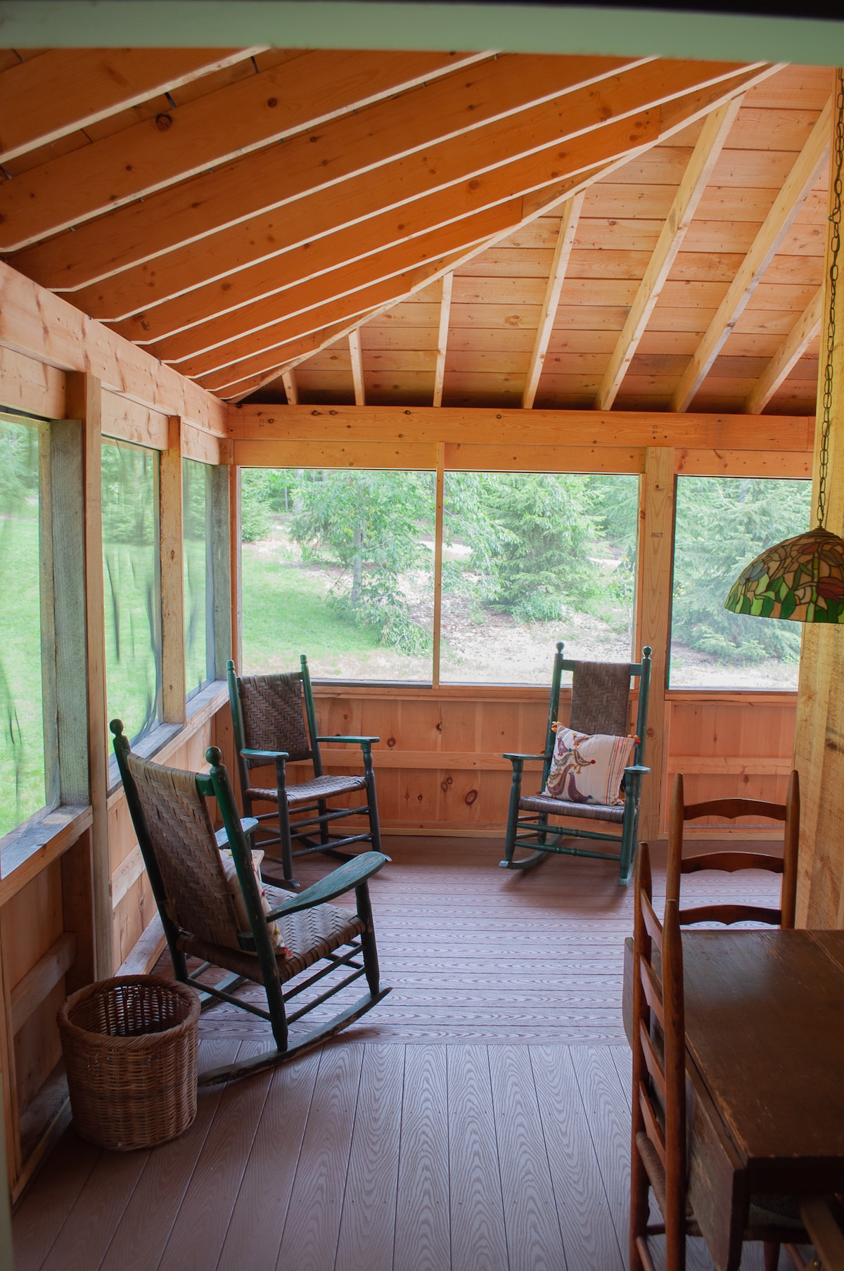 A screened porch features four wooden rocking chairs, inviting relaxation. Natural light filters through large windows, displaying the green space outside. The wooden walls and ceiling provide a warm, rustic feel, while a small basket and dining table enhance the cozy atmosphere.